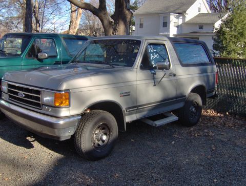 1996 Ford Bronco - Two Ford Broncos are parked outside a house on a sunny day. The gray Bronco has a sign in the window.
