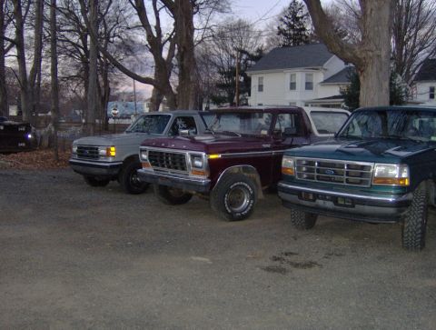 1996 Ford Bronco - Three Ford trucks, including a 1996 Bronco, are parked in a gravel lot near houses.