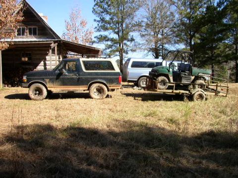 1995 Ford Bronco - A 1995 Ford Bronco is parked in a grassy area next to a log cabin, with a utility vehicle on a trailer behind it.