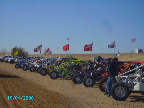 1990 Ford Bronco - A row of dune buggies are parked on a sandy terrain under a clear blue sky, with various flags flying above.
