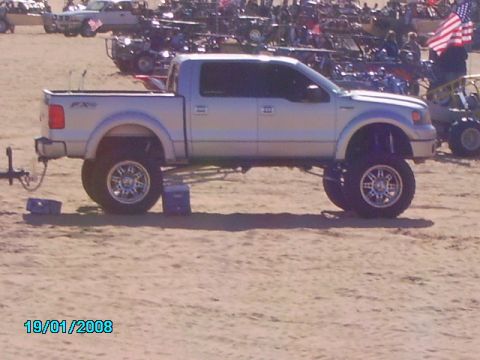 1990 Ford Bronco - Lifted silver Ford F-150 pickup truck with custom wheels on sand, with other vehicles in the background.