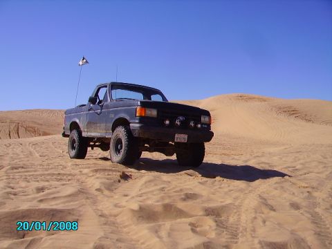 1990 Ford Bronco - This black pickup truck, likely a 1990 model, navigates sandy terrain under a clear blue sky, a pirate flag fluttering in the breeze.
