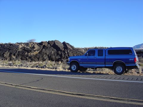 1996 Ford F-250 - A blue pickup truck with a camper shell is parked on the side of a road next to a rocky landscape.