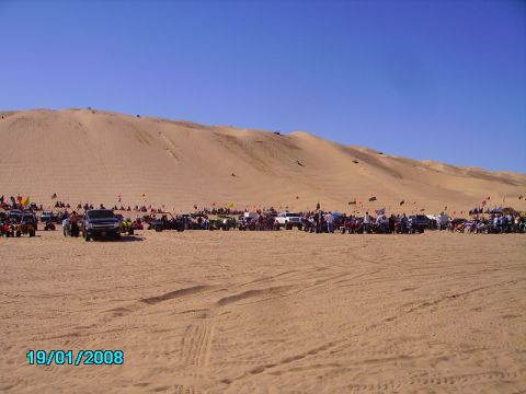 1990 Ford Bronco - A large group of people and various vehicles are gathered at the base of sand dunes under a clear blue sky.
