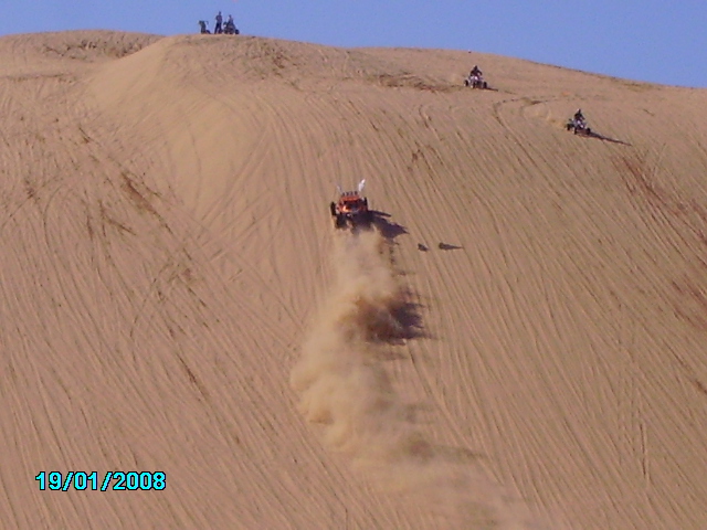 Orange dune buggy descending a sand dune, kicking up dust. Three ATVs are visible on the dune crest
