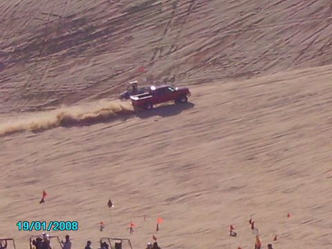 1990 Ford Bronco - A red pickup truck drives across a sandy dune, leaving a trail of dust in its wake.