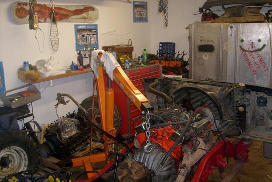 Garage scene with engine hoist over a disassembled vehicle engine and parts. A large metal cabinet with a smiley face is in the background