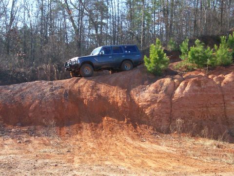 1993 Ford Bronco - A dark blue SUV navigates a steep, red dirt incline surrounded by trees.