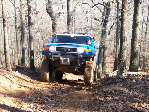 1993 Ford Bronco - A blue Toyota FJ Cruiser navigates a wooded trail with fallen leaves on the ground.