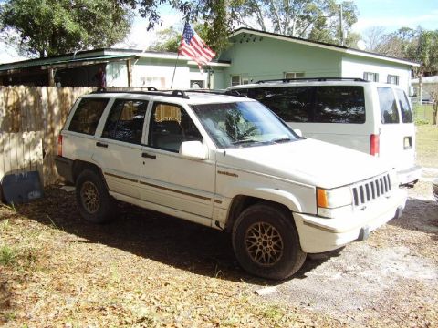 2004 Jeep TJ - A white Jeep Grand Cherokee is parked in front of a building with a white van behind it.