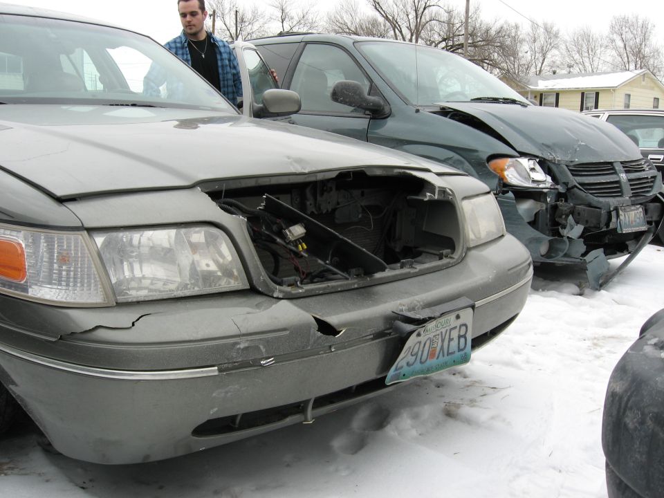 Wrecked gold Ford Crown Victoria with grille missing in a snowy salvage yard