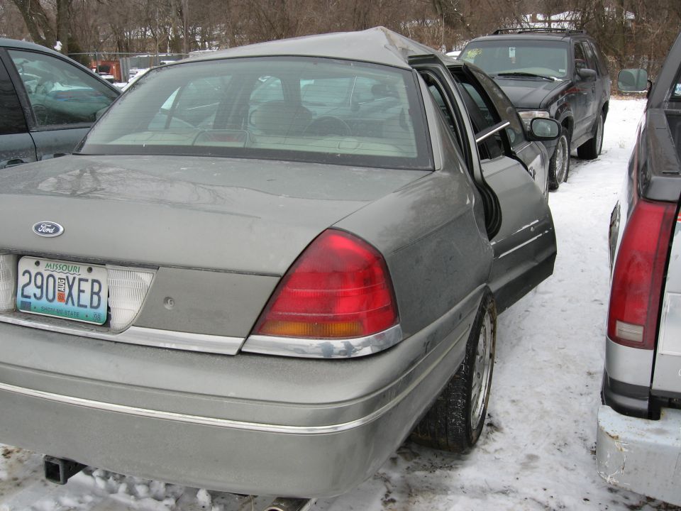 Wrecked silver-green Ford Crown Victoria rear quarter view in snowy salvage lot, Missouri plate