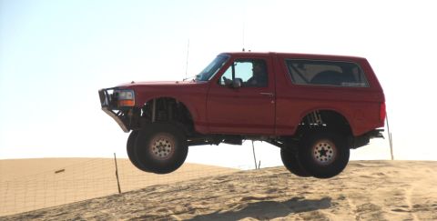 1984 Ford Bronco - A red Ford Bronco soars through the air over a sand dune, its lifted suspension and off-road capabilities.