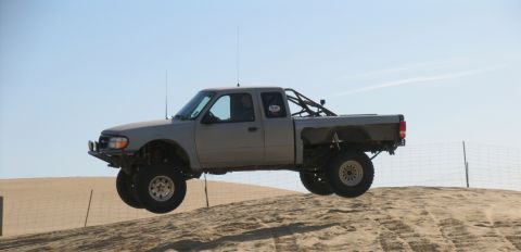 1984 Ford Bronco - A modified gray Ford Ranger truck is captured mid-air during a jump over a sand dune with a clear blue sky in the background.