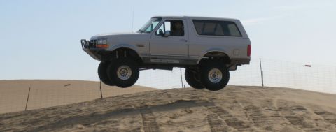 1984 Ford Bronco - Silver Ford Bronco airborne over a sand dune with a clear sky in the background.