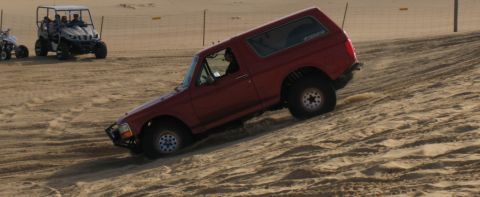 1984 Ford Bronco - A red SUV navigates a sandy incline, with other recreational vehicles visible in the distance.