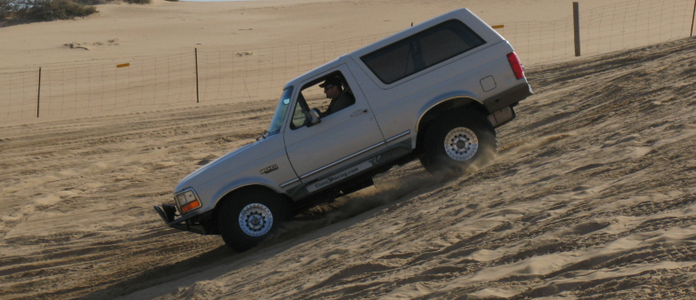 Silver Ford Bronco driving up a sand dune, driver visible
