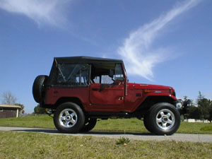 1991 Ford Bronco - This red convertible SUV is shown from a 3/4 front angle under a blue sky with wispy clouds. The Supermotors logo is visible on the lower r…