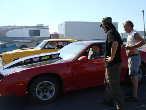 1985 Chevrolet Camaro - Three men stand around a red 1985 Chevrolet Camaro with a large hood scoop and racing stickers.