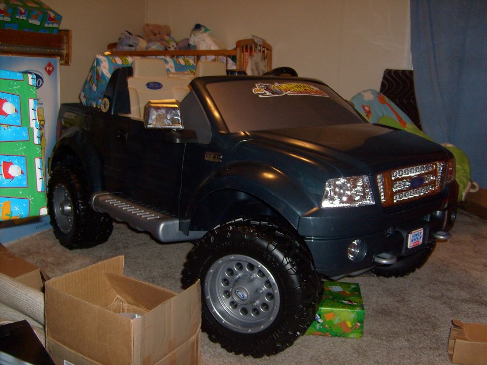 Dark green Power Wheels Ford F-150 ride-on toy truck on carpet indoors, surrounded by boxes and wrapped gifts