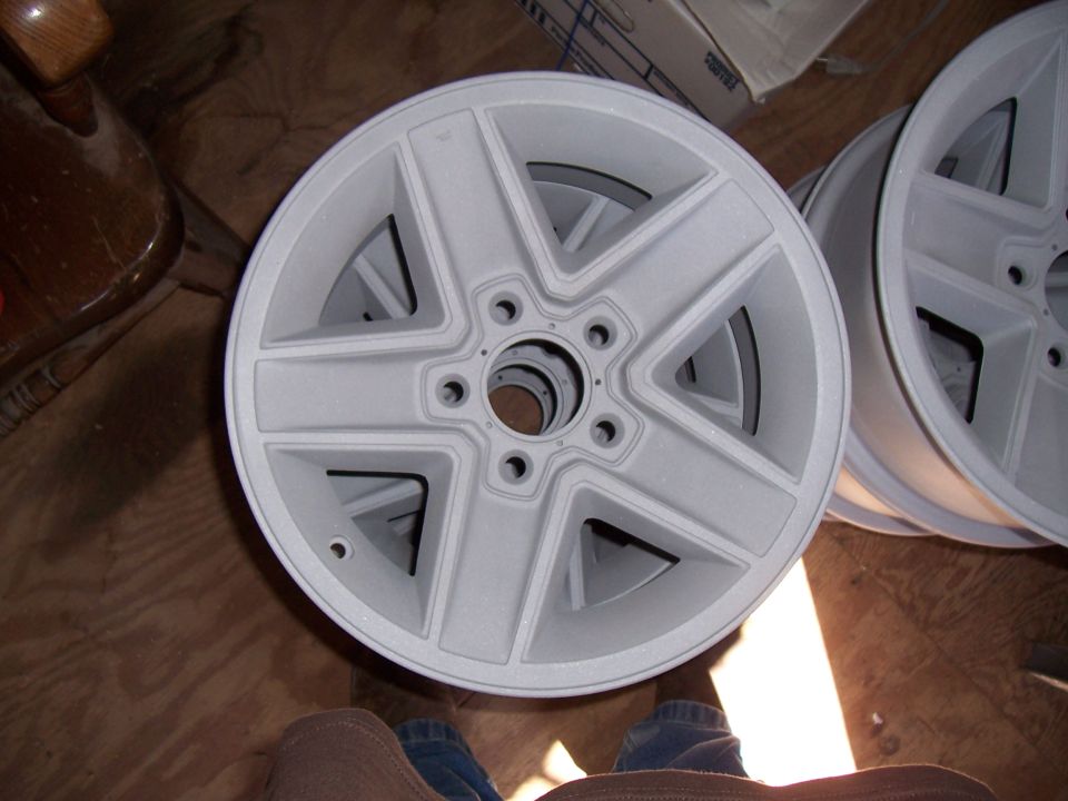 Pair of unfinished five-spoke wheels, one in foreground, stacked on wooden floor