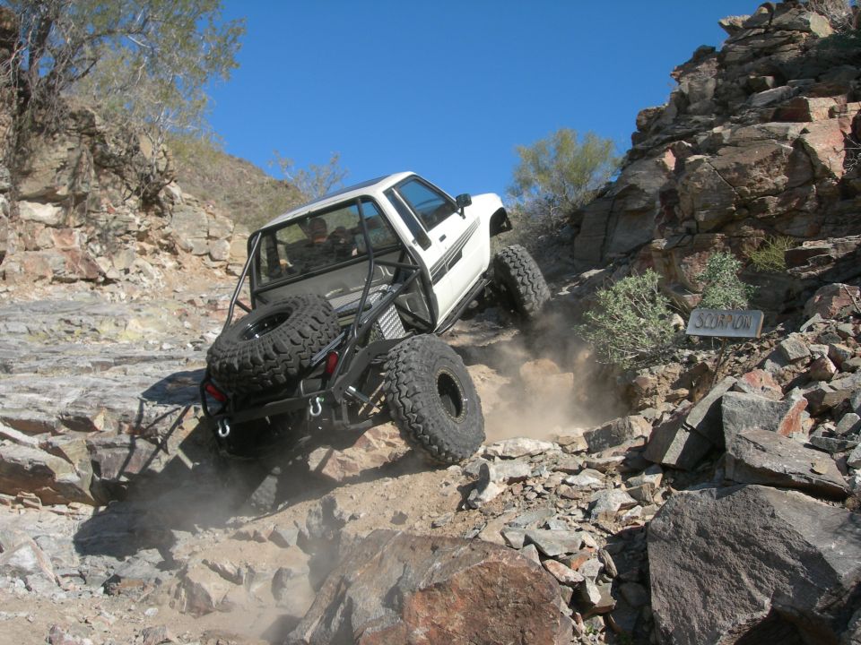White pickup truck with roll cage and spare tire on back climbing a steep, rocky hill. Sign says 'SCORPION'