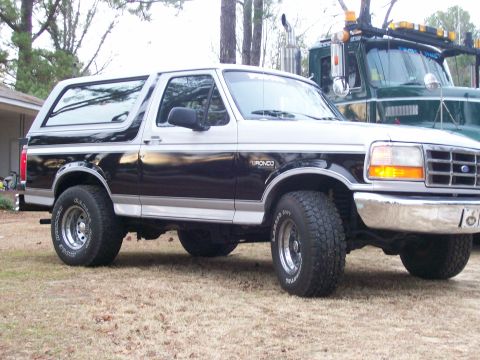 1996 Ford Bronco - A two-tone Ford Bronco is parked in front of a large green tow truck.