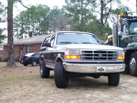 1996 Ford Bronco - A silver and brown Ford Bronco is parked in a yard next to a large truck and a house.
