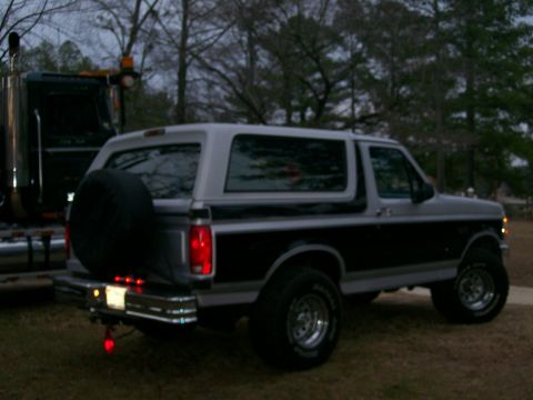 1996 Ford Bronco - A two-tone Ford Bronco is parked outdoors next to a large truck in the evening.