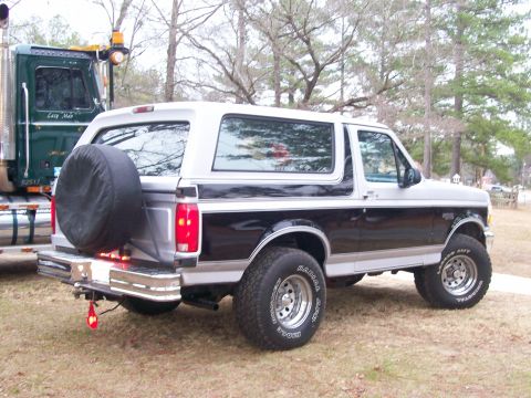 1996 Ford Bronco - A two-tone Ford Bronco is parked next to a large semi-truck, with a spare tire mounted on the back.