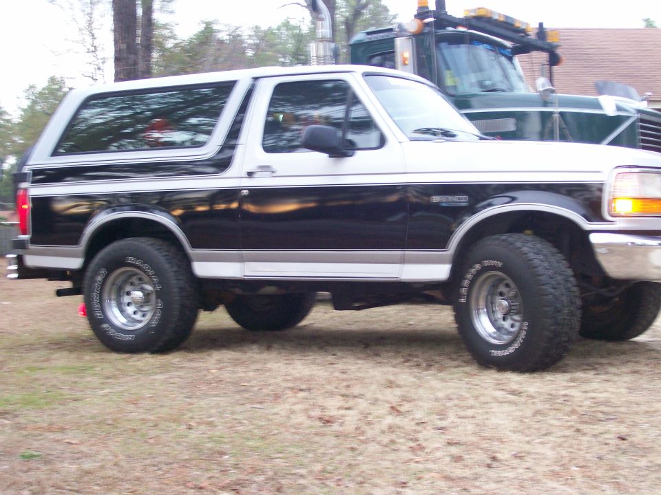 Two-tone black and silver Ford Bronco parked on grass, with a green tow truck behind it