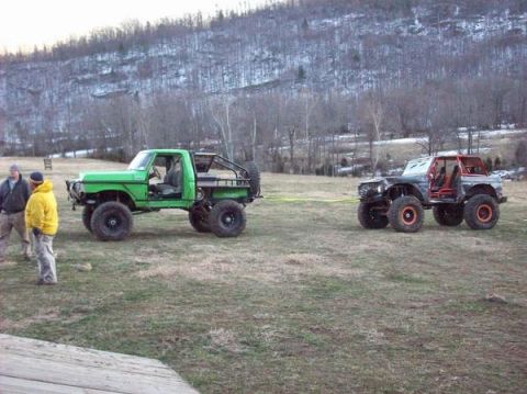 1978 Ford Bronco - Two heavily modified trucks, one green pickup and one black SUV, are parked on a grassy field with a wooded hillside in the background.