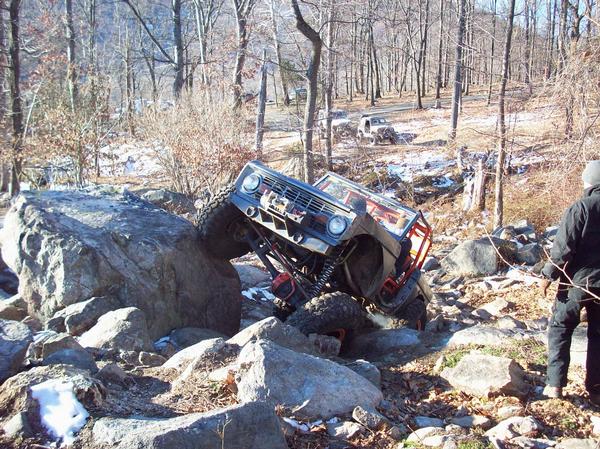 1978 Ford Bronco climbing over large rocks on a wooded trail, another vehicle visible in background