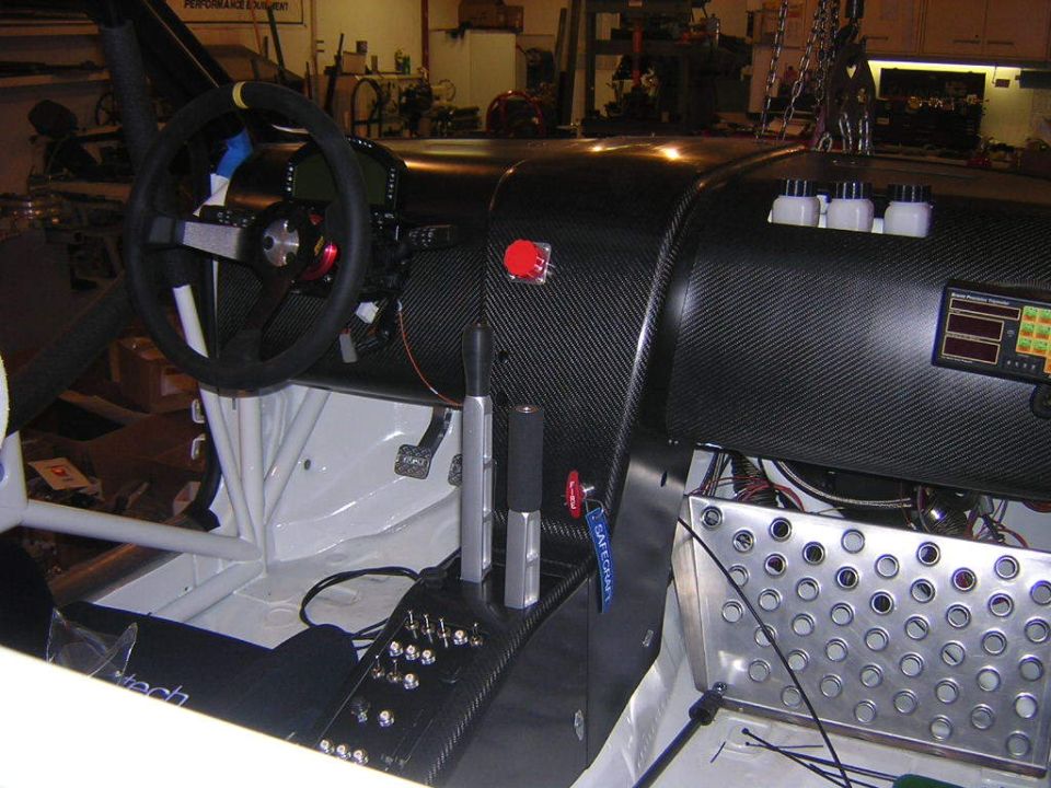 Racing car interior showing carbon fiber dashboard, steering wheel, shifter, and pedals. A digital display is visible