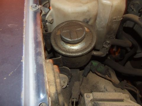 1990 Ford Bronco - Close-up of a power steering fluid reservoir in an engine bay, showing a T-shaped cap and surrounding components.