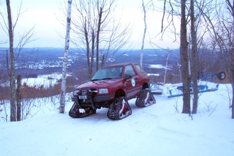 1987 Suzuki Samurai - A red SUV equipped with snow tracks navigates a snow-covered terrain with a scenic view in the background.