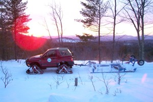 1987 Suzuki Samurai - A red SUV equipped with snow tracks is seen towing a trailer through a snow-covered area during sunset.