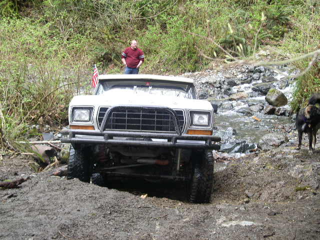 White SUV with brush guard driving on a muddy trail next to a stream. Man stands on top of the vehicle