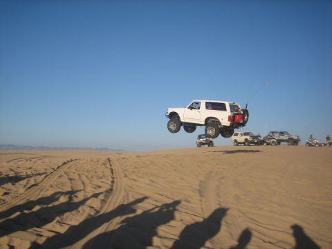 1995 Ford Bronco - A white 1995 Ford Bronco is captured mid-air while driving over sand dunes, with other vehicles visible in the distance.