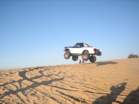 1995 Ford Bronco - A modified 1995 Ford Bronco is airborne over a sand dune with another vehicle in the background.