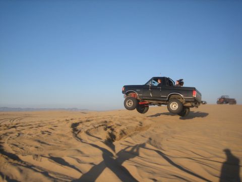 1995 Ford Bronco - A black 1995 Ford Bronco is captured mid-air as it jumps over a sand dune with another vehicle in the background.