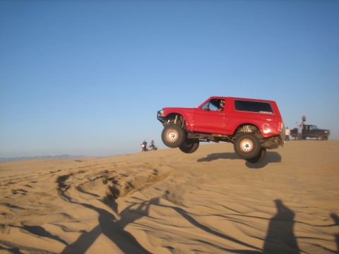 1995 Ford Bronco - Red 1995 Ford Bronco airborne over a sand dune, with another vehicle visible in the distance.