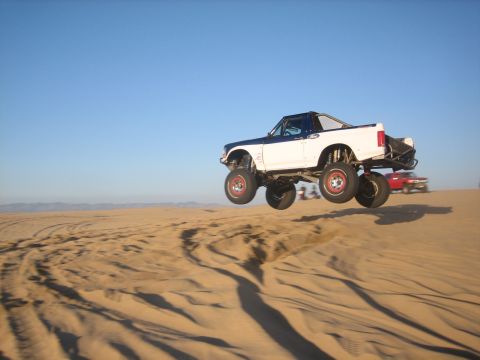1995 Ford Bronco - A modified pickup truck soars through the air over sand dunes under a clear blue sky.