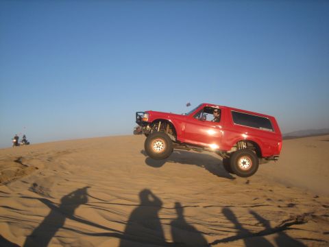 1995 Ford Bronco - A red Ford Bronco is captured mid-air as it jumps over a sand dune under a clear blue sky.