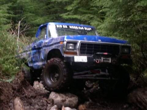 1979 Ford Bronco - A blue 1979 Ford Bronco navigates a challenging off-road course with dense foliage in the background.