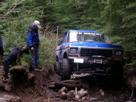 1979 Ford Bronco - A blue 1979 Ford Bronco navigates a challenging off-road trail in a wooded area.