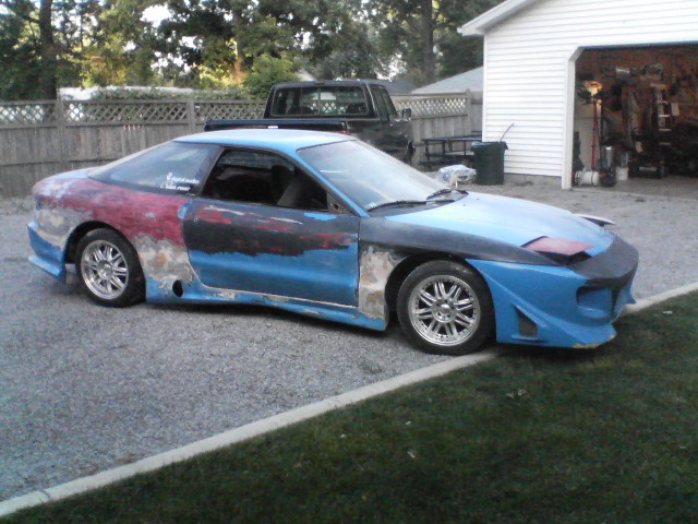 Blue Ford Probe with primer and red paint patches, undergoing body modifications. Hood is black, front bumper is custom