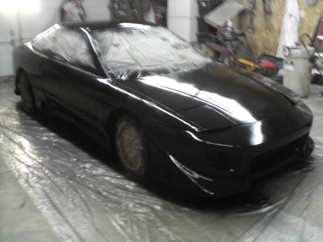 Black car covered in plastic sheeting, parked on a tarp in a garage
