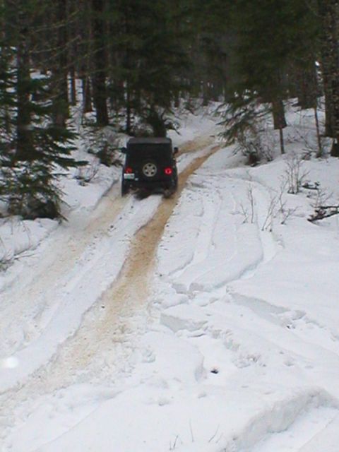 2005 Jeep Wrangler - Rear view of a dark Jeep Wrangler with a rear-mounted spare tire and brake lights lit, driving away up a snow-covered forest road with a mu…