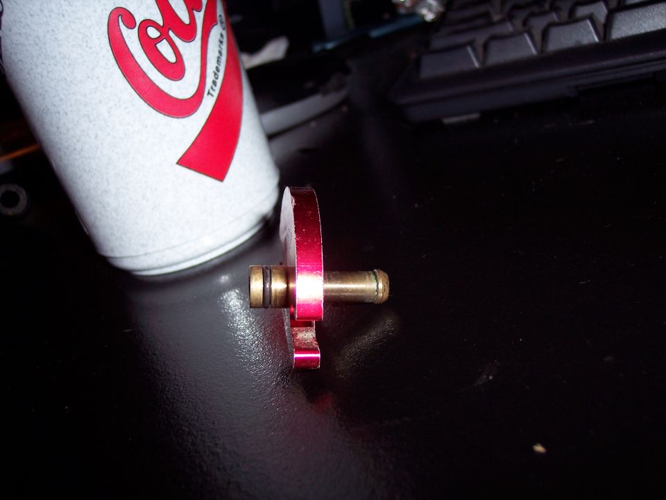 Red metal fuel rail adapter with brass fittings next to a Coca-Cola can